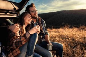 Casal feliz sentado no porta malas de um carro.