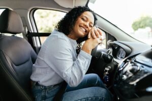 Mulher vestindo uma camiseta de manga longa, no interior de um veículo, debruçada no volante do carro, com os olhos fechados e sorrindo.